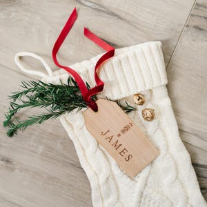 以下が含まれることがあります： 白いニットのクリスマスストッキングに、木製のタグが付けられています。タグには「JAMES」と小さなクリスマスツリーのデザインが刻まれています。赤いリボンでストッキングに結ばれています。