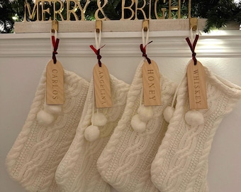May include: Four cream-colored cable knit Christmas stockings hang from a wooden holder with the gold text MERRY & BRIGHT. Each stocking features a wooden name tag labeled Carlos, Angelina, Honey, or Whiskey, tied with a red ribbon.