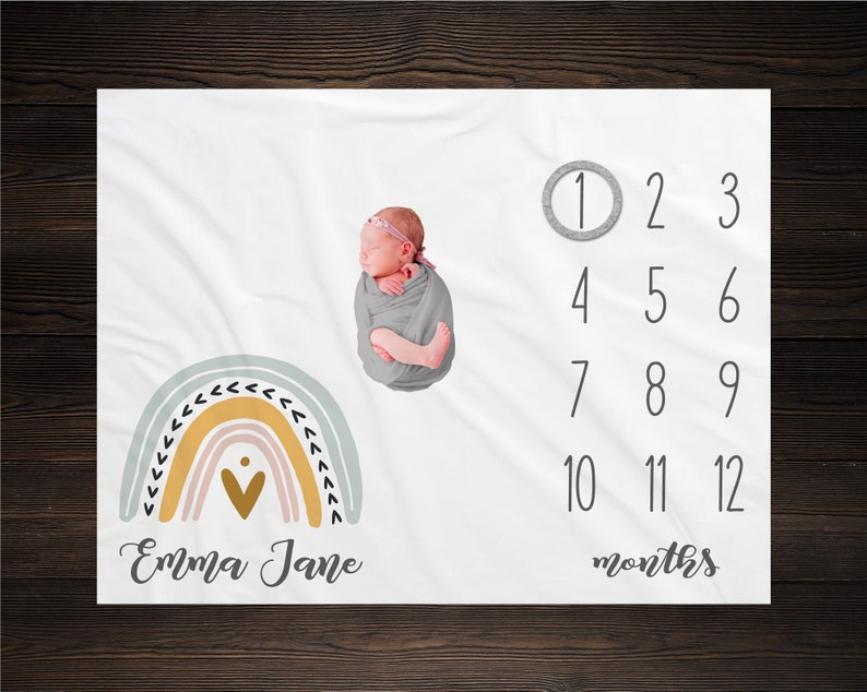 May include: A white baby blanket with a rainbow design and a heart in the center. The blanket also has a number chart for tracking a baby's growth, with the text "Emma Jane" and "months" printed on the blanket.