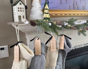 May include: Four knit Christmas stockings in cream and gray hanging from a mantel, each featuring a wooden tag engraved with the names Lucy, Benson, Sarah, and Mike, tied with black ribbon. The stockings have a textured cable knit pattern.