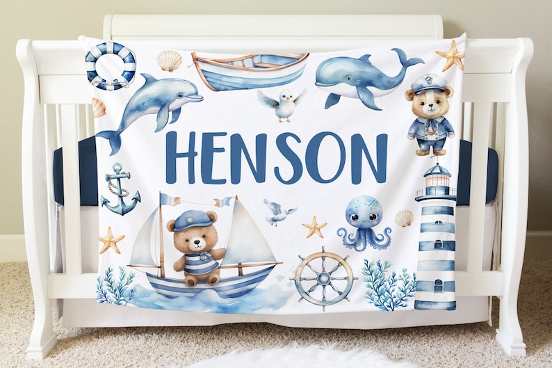 May include: A white baby blanket with a nautical theme. It features a blue and white striped lighthouse, a blue whale, a blue and white sailboat with a teddy bear wearing a captain's hat, a blue octopus, and the name "HENSON" in blue letters.
