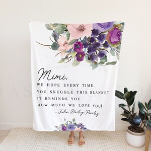 Puede incluir: Una manta blanca con un dise&ntilde;o floral en tonos de morado, rosa y verde. La manta tiene un mensaje personalizado que dice: "Mimi, Esperamos que cada vez que te acurruques en esta manta, te recuerde cu&aacute;nto te queremos! - Jalie Hailey Peisley".