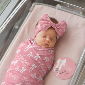 May include: A dusty pink swaddle blanket and matching headband with a large bow, both featuring a repeating white pattern of bows and the name Amber Olivia in script. A circular pink birth announcement card displays the name Amber Olivia.