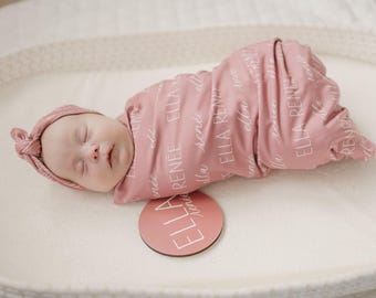 May include: A sleeping baby wrapped in a pink swaddle blanket and matching headband. The blanket and headband have the name "Ella Renee" printed on them. A pink wooden circle with the name "ELLA" is next to the baby.