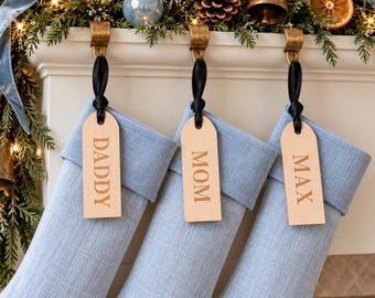 Op de afbeelding: Drie lichtblauwe stoffen kerstsokken hangen aan goudkleurige metalen haken. Elke sok is voorzien van een glad, lichtgekleurd houten label dat met een zwart lint is vastgemaakt en gegraveerd is met de namen DADDY, MOM en MAX.