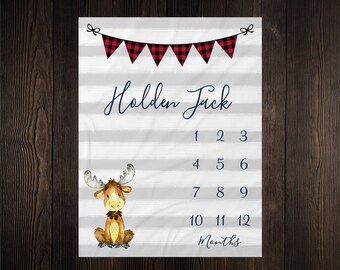 Op de afbeelding: Een wit en grijs baby-mijlpaal deken met rood en zwart buffalo plaid bunting. Het deken heeft een eland illustratie en de nummers 1 tot en met 12 erop gedrukt. De tekst "Holden Jack" en "Maanden" is op het deken gedrukt.