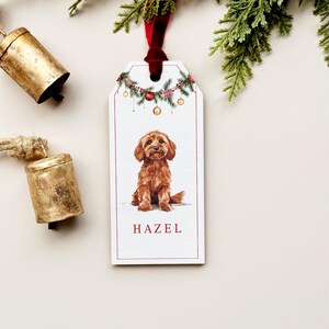 May include: A white rectangular gift tag with a red border features a watercolor illustration of a brown dog named Hazel. The tag is decorated with Christmas garland and a red velvet ribbon.