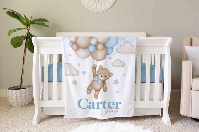 May include: A white baby blanket with a brown teddy bear holding onto blue and brown balloons. The blanket has a blue and white cloud design and gold stars. The text "Carter James" is printed in blue at the bottom of the blanket.