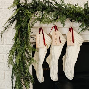 Puede incluir: Tres calcetines de punto blanco con etiquetas de madera colgando de una repisa de chimenea. Las etiquetas están etiquetadas como "Brooks", "Mommy" y "Dad".