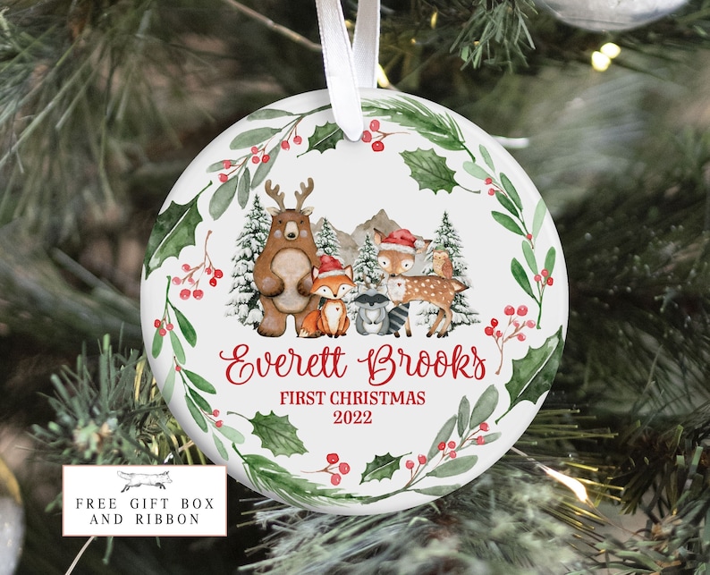 Op de afbeelding: Ronde witte kerstbal met een aquarelontwerp van bosdieren, waaronder een beer, een vos, een hert en een wasbeer. De kerstbal is versierd met hulst en bessen. De tekst "Everett Brooks First Christmas 2022" staat op de kerstbal gedrukt.