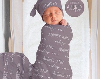 Può includere: Un neonato avvolto in una coperta grigia e un cappello abbinato, entrambi stampati con il nome "Aubrey Ann" in un motivo ripetuto. Il bambino è sdraiato su un asciugamano bianco all'interno di una culla trasparente.