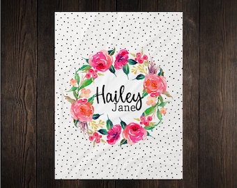 May include: A white baby blanket with a black polka dot pattern. A pink floral wreath with the name "Hailey Jane" in black script is in the center.