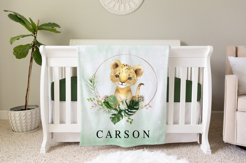 May include: A soft green and white baby blanket with a watercolor illustration of a lion cub in a gold circle surrounded by green leaves. The blanket has the name "CARSON" printed in black at the bottom.