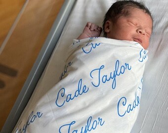 Może przedstawiać: Biały otulacz z imieniem "Cade Taylor" napisanym niebieskim pismem odręcznym. Noworodek jest owinięty w otulacz.