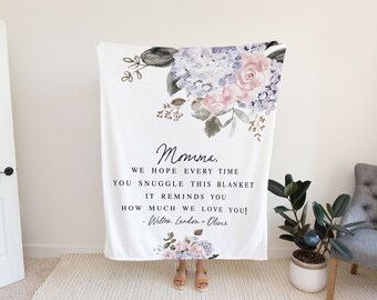 Pode incluir: Um cobertor branco com um design floral em tons de rosa, roxo e verde. O cobertor tem uma mensagem que diz: "Mamãe, esperamos que cada vez que você se aconchegue neste cobertor, ele lhe lembre o quanto nós a amamos! - Willow, London + Oliver"