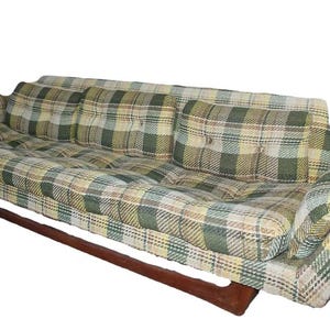 May include: A vintage plaid sofa with a wooden frame. The sofa features a green, yellow, and beige plaid pattern on the cushions and back. The sofa has a low profile and a mid-century modern design. The sofa is approximately 80 inches long.