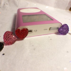 May include: Five heart-shaped charms with rhinestones in different colors: black, pink, red, purple, and white. The charms are sitting on a white surface next to a pink iPod.