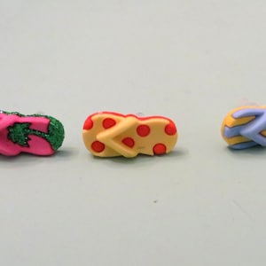 May include: Three colorful flip-flop charms with different designs. The first charm is pink with green glitter and a green palm tree design. The second charm is yellow with red polka dots. The third charm is blue with yellow stripes.