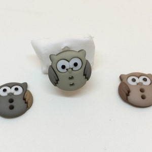 May include: Three owl-shaped buttons in different colors: gray, brown, and green. Each button has white eyes and two black dots for a nose and mouth.