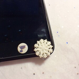 May include: A black phone case with a white snowflake charm and a St. Louis Blues logo sticker. The snowflake is detailed and three-dimensional. The case has a glossy finish.
