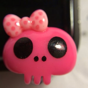 May include: A pink skull-shaped charm with large black eyes and a small bow with pink polka dots. The charm has a skull-like jawline and two small dots for nostrils. The bow is positioned on top of the skull.