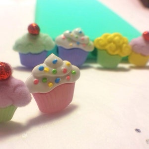 May include: A collection of six colorful cupcake-shaped buttons with white frosting and sprinkles. The buttons are made of plastic and have a shiny finish.