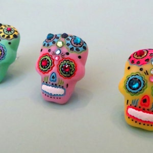 May include: Three colorful sugar skull ornaments with rhinestones. The skulls are painted in green, pink, and yellow with black details and colorful flowers.