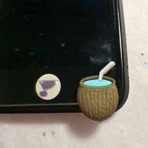 May include: A brown coconut shaped phone charm with a blue liquid inside and a white straw sticking out of the top. The charm is attached to a black phone case with a blue and white hockey team logo sticker.