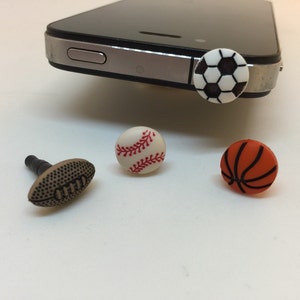 May include: Four different phone charms, each featuring a different sport: a baseball, a basketball, a football, and a soccer ball. The charms are made of plastic and have a white background with black and red details.