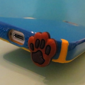 May include: A blue phone case with a yellow stripe and a brown paw print charm.