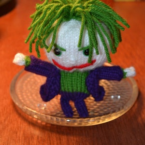 May include: A crocheted toy figure of the Joker from Batman, with green hair, a white face, a purple and green outfit, and a red smile. The toy is sitting on a clear glass plate.