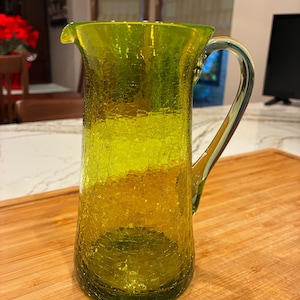 May include: A green glass pitcher with a crackle finish. The pitcher has a spout and a curved handle. The pitcher is sitting on a wooden cutting board. The pitcher is approximately 8 inches tall.