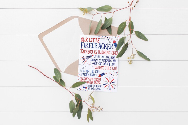 May include: A festive invitation for a first birthday celebration. The card features red, white, and blue text and illustrations, including fireworks and a rocket. The invitation is placed inside a light brown envelope, with greenery accents.
