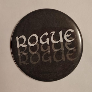 May include: A circular pin-back button with a black background. The word ROGUE is printed in a white, stylized serif font across the upper half. Below the white text, the word ROGUE is repeated twice in a faint, dark gray, semi-transparent font.