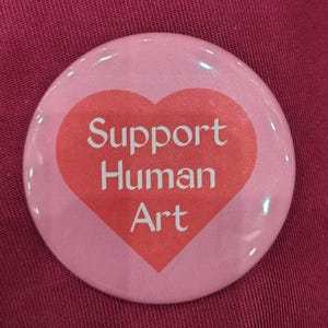 May include: A round pinback button featuring a light pink background and a large, centered coral-colored heart. Inside the heart, the white text reads Support Human Art in a clean, sans-serif font. The button has a glossy, reflective finish.
