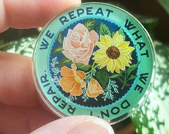 May include: A round, blue and green pin with a floral design and the text "We repeat what we don't repair."