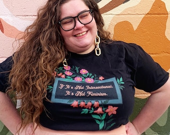 Może przedstawiać: Osoba w czarnym t-shircie z napisem "If It's Not Intersectional, It's Not Feminism." i różową tiulową spódnicą, stojąca przed ścianą z muralem kwiatowym.