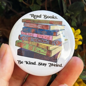 Puede incluir: Un botón blanco con una pila de libros coloridos y el texto "Read Books. Be Kind. Stay Weird."