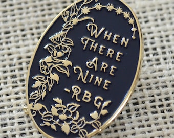 May include: A gold-colored enamel pin with a black background and a floral design. The pin features the quote "When there are nine - RBG".