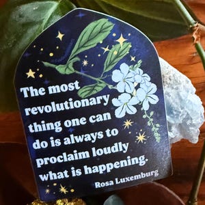 May include: A sticker with a dark blue background, white flowers, and gold stars. The text reads, "The most revolutionary thing one can do is always to proclaim loudly what is happening." Quote by Rosa Luxemburg.
