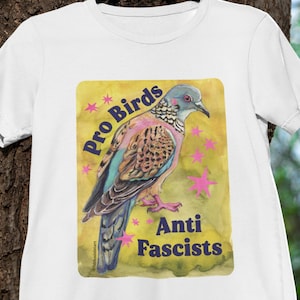May include: White t-shirt featuring a colorful illustration of a bird with the text "Pro Birds Anti Fascists". The bird has brown, blue, and pink feathers. The design is on a yellow background with pink star accents.
