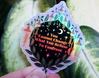 Op de afbeelding: Een holografische sticker met een zwarte achtergrond en een citaat dat luidt: "Je kunt niet veranderen wat je weigert te confronteren." De sticker is omgeven door een bloemmotief met groene bladeren en roze bloemen.