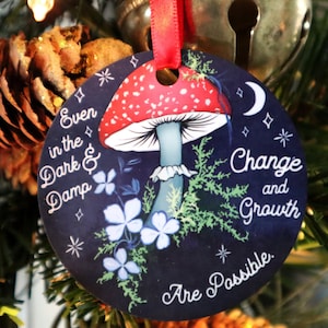 May include: Round Christmas ornament with a red and white mushroom design on a dark blue background. The ornament has the words "Even in the Dark & Damp" and "Change and Growth Are Possible." A red ribbon is attached for hanging.