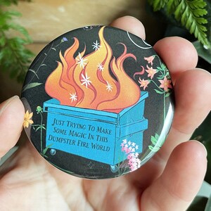 May include: A round button with a black background and a blue dumpster on fire with orange flames and white stars. The text on the button reads "Just trying to make some magic in this dumpster fire world."