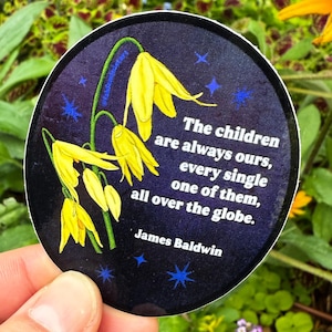 May include: A circular sticker with a dark blue backdrop, adorned with yellow flowers and blue stars. The sticker bears the quote "The children are always ours, every single one of them, all over the globe." by James Baldwin.