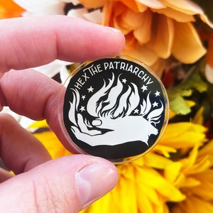 May include: A black and white round pin with a hand holding a flame with stars around it. The text on the pin reads "Hex the Patriarchy".