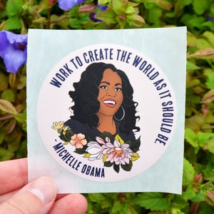 Pode incluir: Um adesivo redondo com fundo branco e borda azul. O adesivo apresenta um retrato de Michelle Obama com o texto "Trabalhar para criar o mundo como deveria ser." - Michelle Obama.