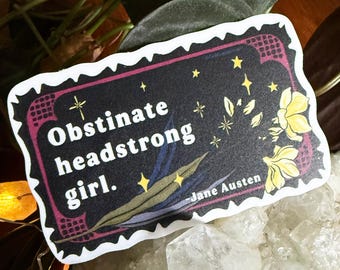 May include: A sticker with the text "Obstinate headstrong girl." in white on a black background, with yellow stars and flowers. The sticker has a white border and is surrounded by green leaves and a white crystal.