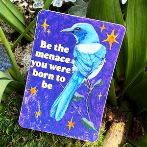 May include: A rectangular sticker with a blue bird illustration and the text "Be the menace you were born to be" on a purple background. The sticker is surrounded by green plants and blue flowers.