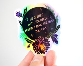 May include: A round sticker with a black background and a floral design. The sticker has a holographic finish and features the text "Be gentle with yourself. You're doing the best you can."
